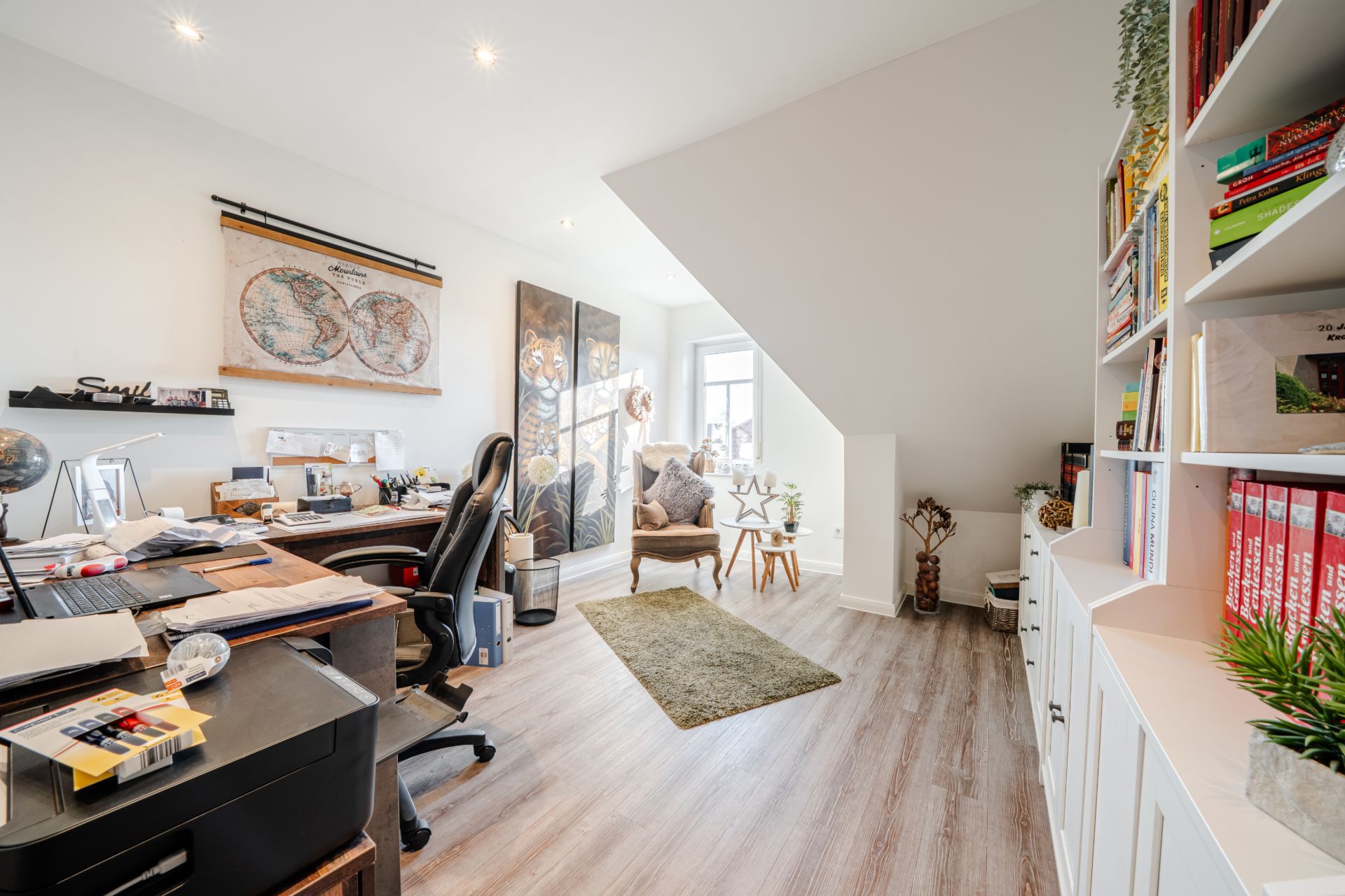 Helles Arbeitszimmer mit Schreibtisch, Regalwand, Stuhl und Sitzbereich am Fenster.