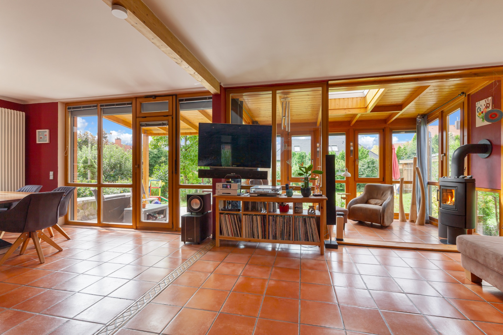 Wohnzimmer mit Terrakottaboden, großen Fenstern, TV, Ofen und Blick auf den Garten.