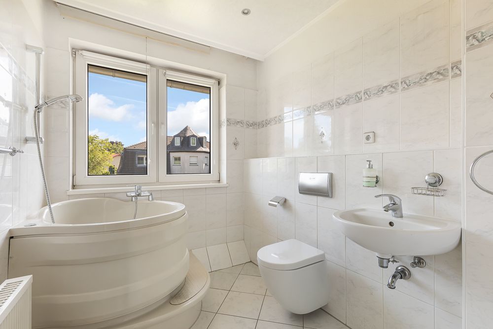 Helles Bad mit Eckbadewanne, Wandtoilette, Waschbecken und Fenster mit Blick nach draußen.