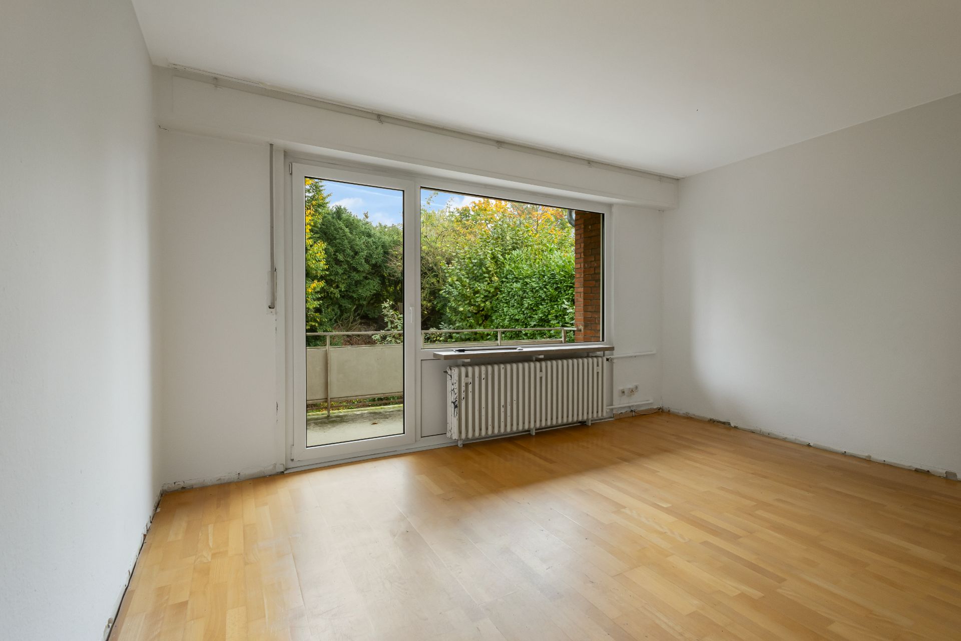 Leeres Zimmer mit Holzfußboden und großem Fenster zur Terrasse mit Blick ins Grüne.