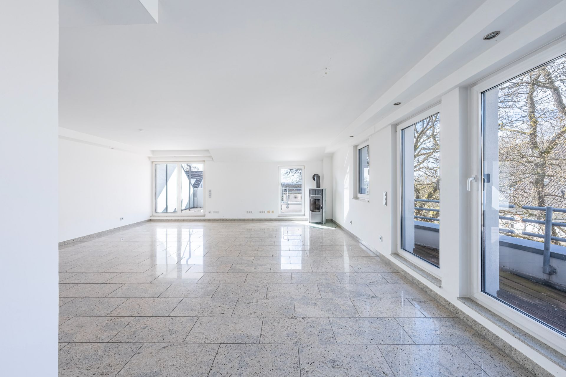 Heller, leerer Raum mit großen Fenstern und Fliesenboden, Zugang zu einem Balkon.