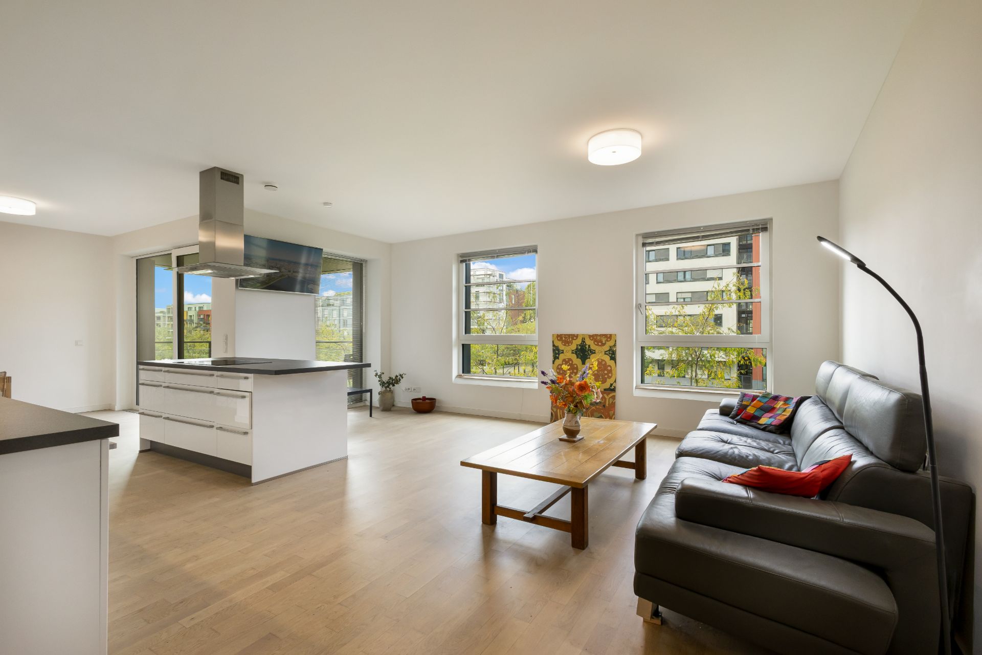 Modernes Wohnzimmer mit grauem Sofa, Holztisch und offener Küche, große Fenster lassen viel Licht hinein.