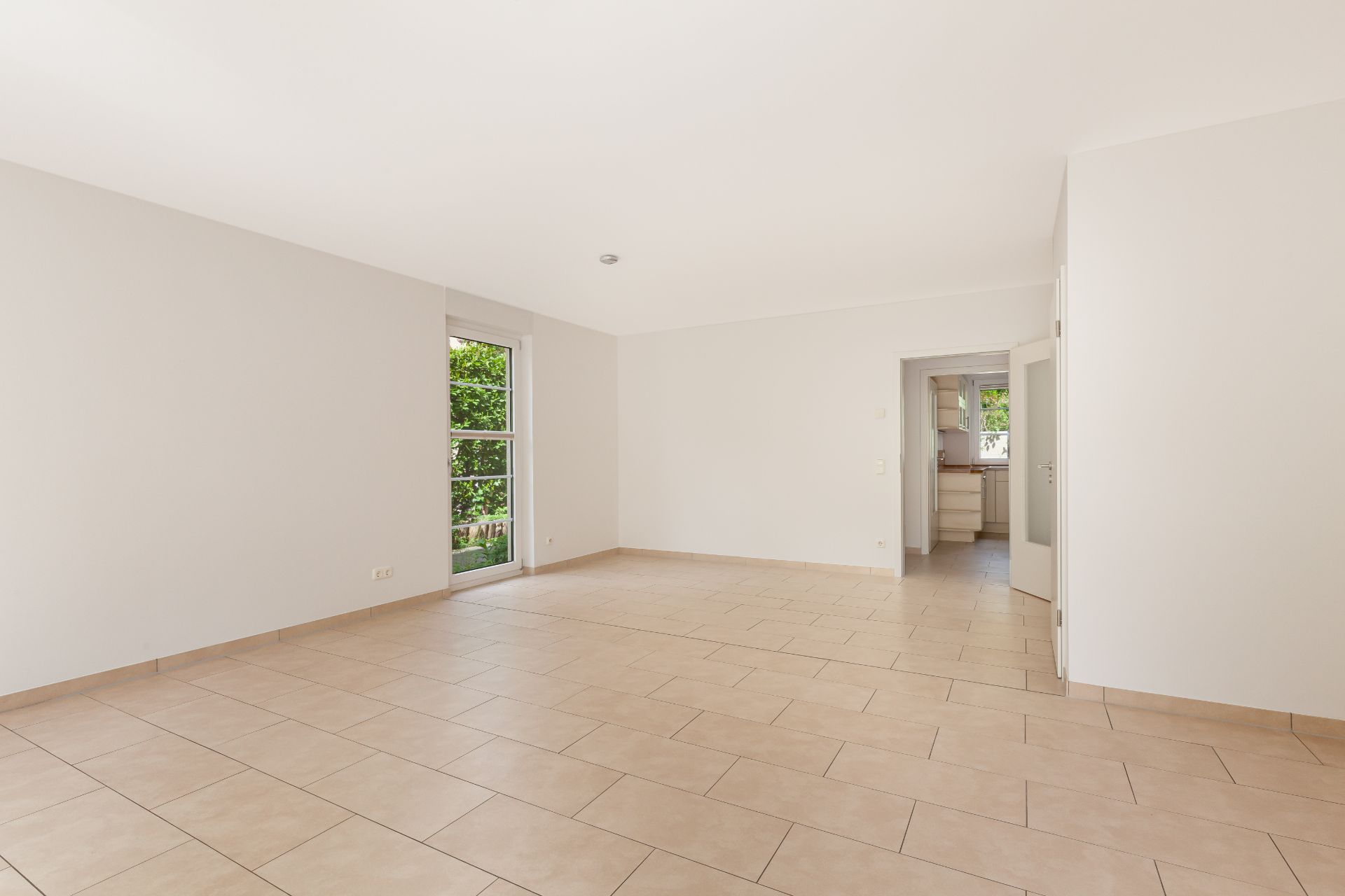 Leerer, heller Raum mit Fliesenboden, großer Fensterfront und Zugang zur Küche.
