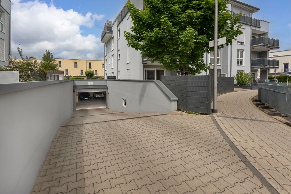 Einfahrt zu einer Tiefgarage neben einem modernen Wohngebäude mit Gehweg und Baum.