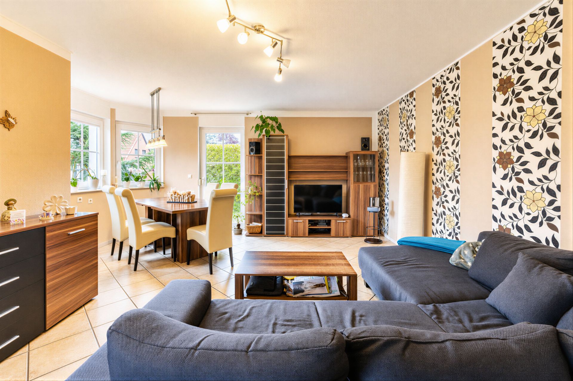 Helles Wohnzimmer mit grauem Sofa, Holztisch, TV-Schrank und Essbereich mit sechs Stühlen.