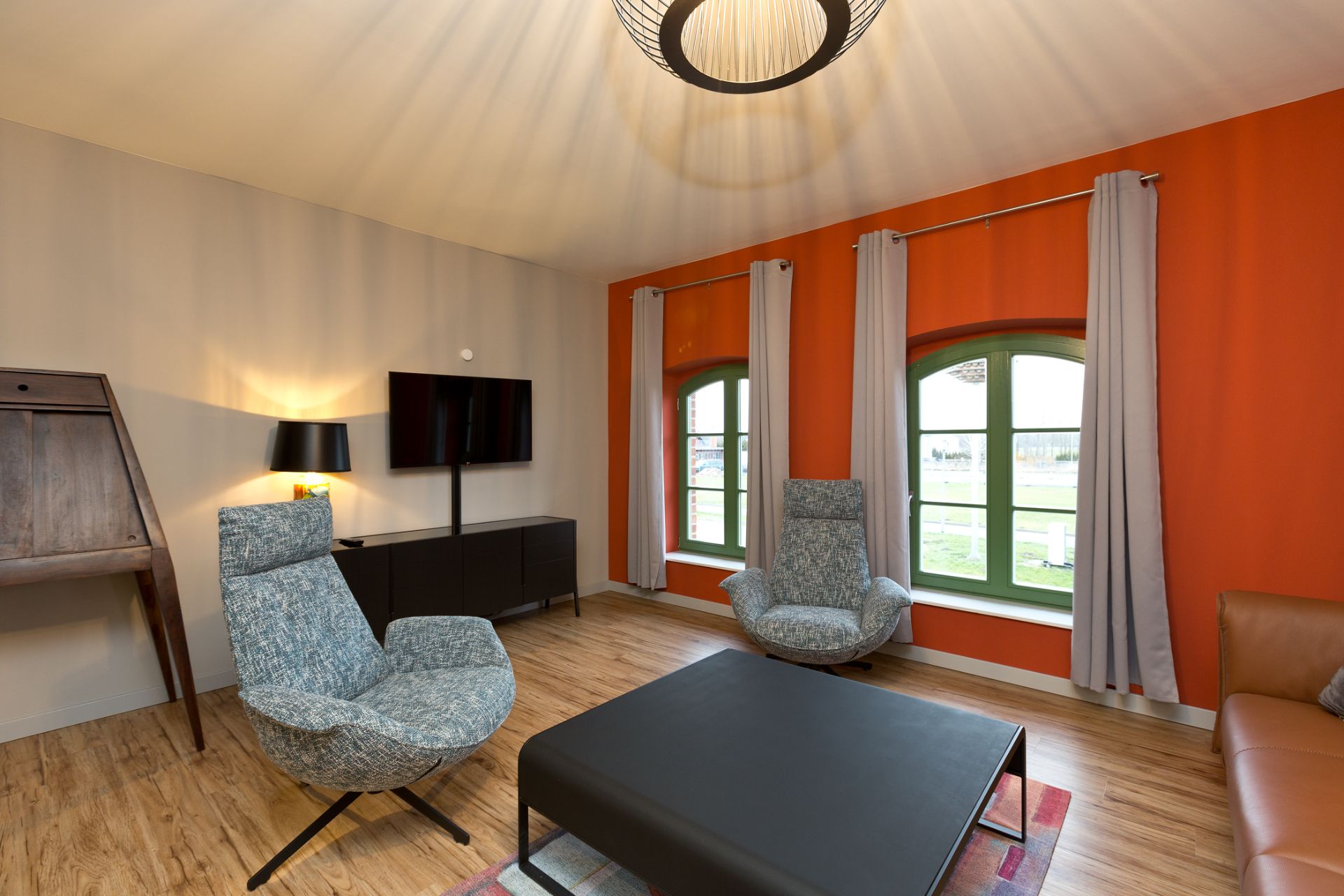 Modernes Wohnzimmer mit zwei Sesseln, Couchtisch, TV und großen Fenstern mit Vorhängen.