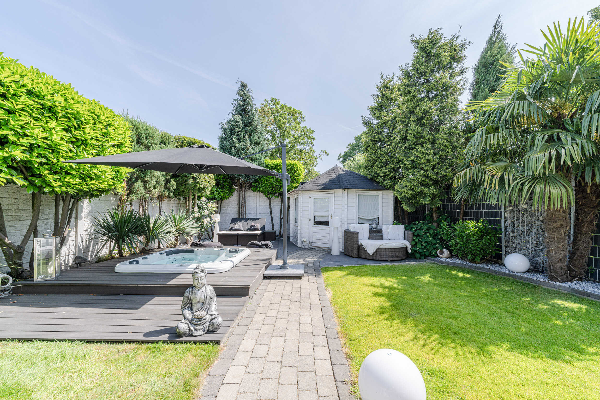 Garten mit Whirlpool, Buddha-Statue, Sitzbereich, Sonnenschirm und Gartenhaus, umgeben von Bäumen.