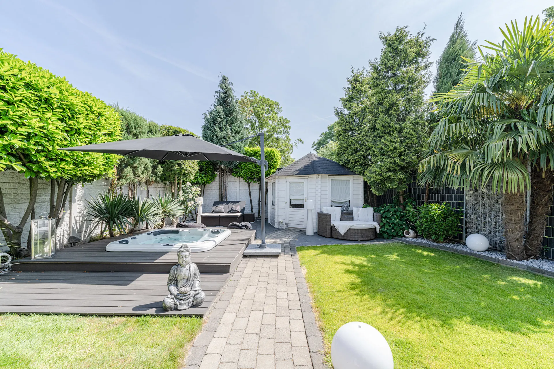 Garten mit Whirlpool, Buddha-Statue, Sitzbereich, Sonnenschirm und Gartenhaus, umgeben von Bäumen.