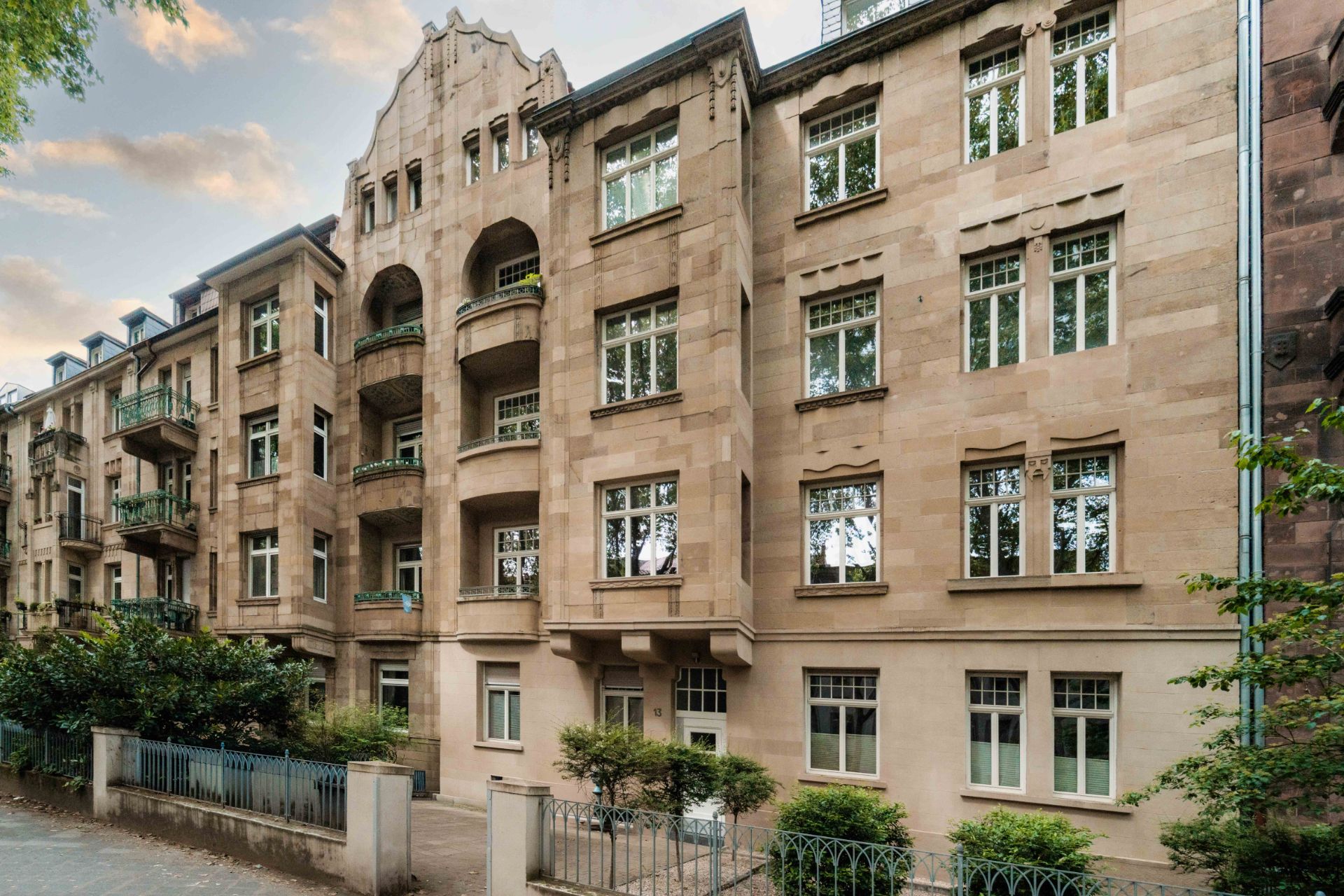 Mehrstöckiges Wohnhaus aus Stein mit Balkonen und großen Fenstern, umgeben von Bäumen und Zaun.