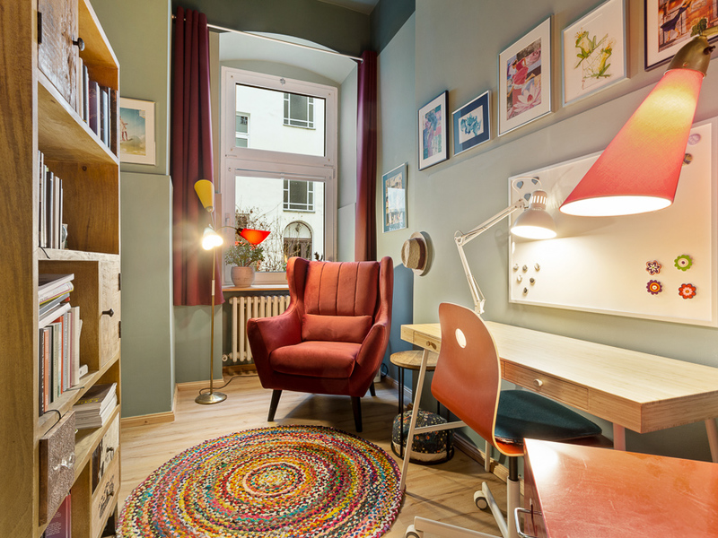 Helles Arbeitszimmer mit rotem Sessel, Schreibtisch, Regal und buntem Teppich.
