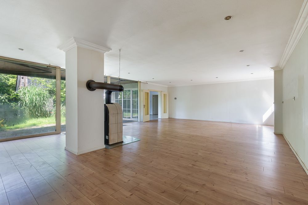 Großes, leeres Wohnzimmer mit Holzboden, Kamin und bodentiefen Fenstern zum Garten.