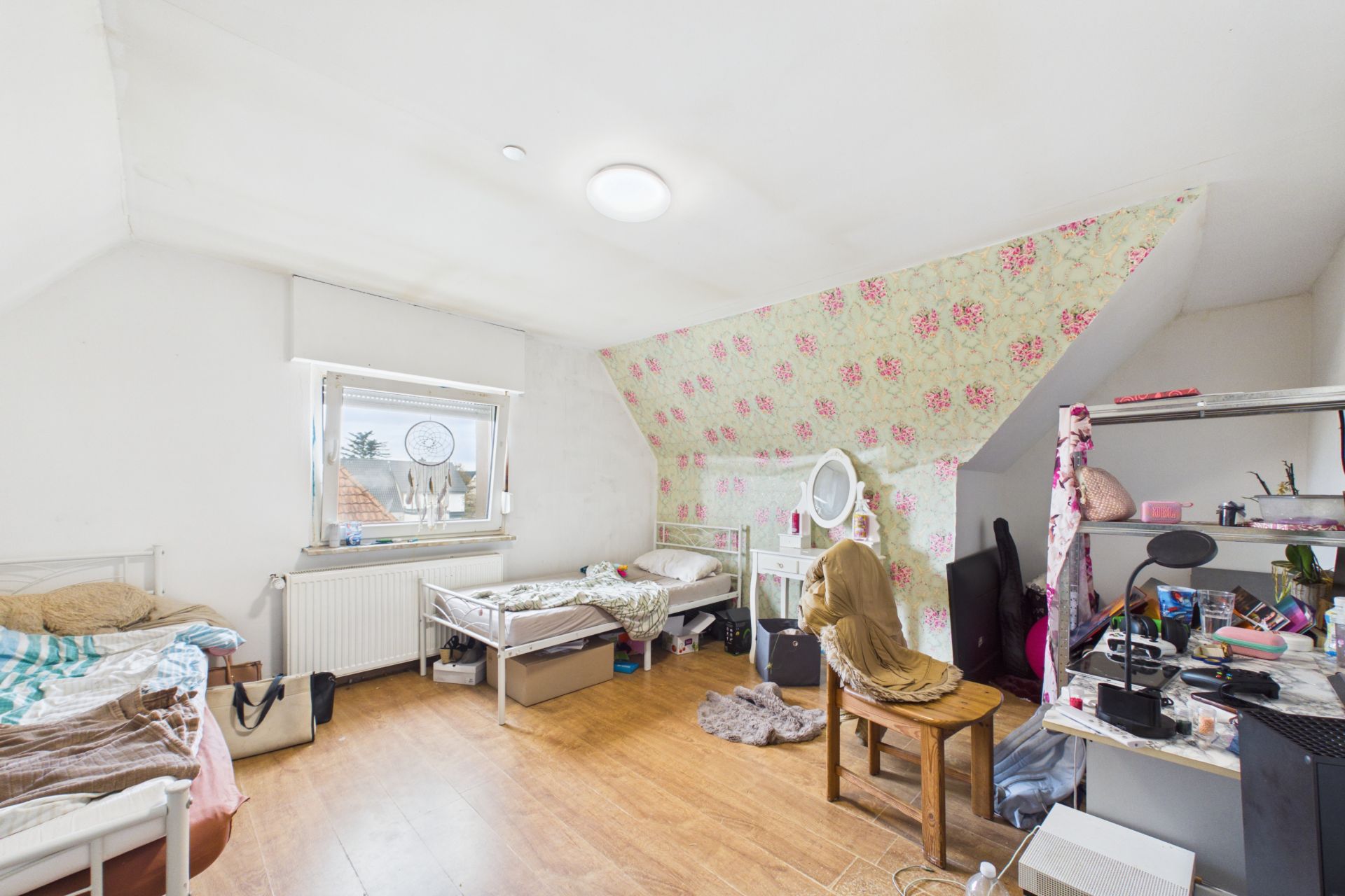 Helles Zimmer mit zwei Betten, Schreibtisch, Stuhl und buntem Blumenmuster an einer Wand.