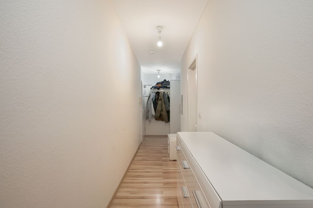 Langer, heller Flur mit Holzboden, an der Seite eine weiße Kommode und Kleider an der Garderobe am Ende.