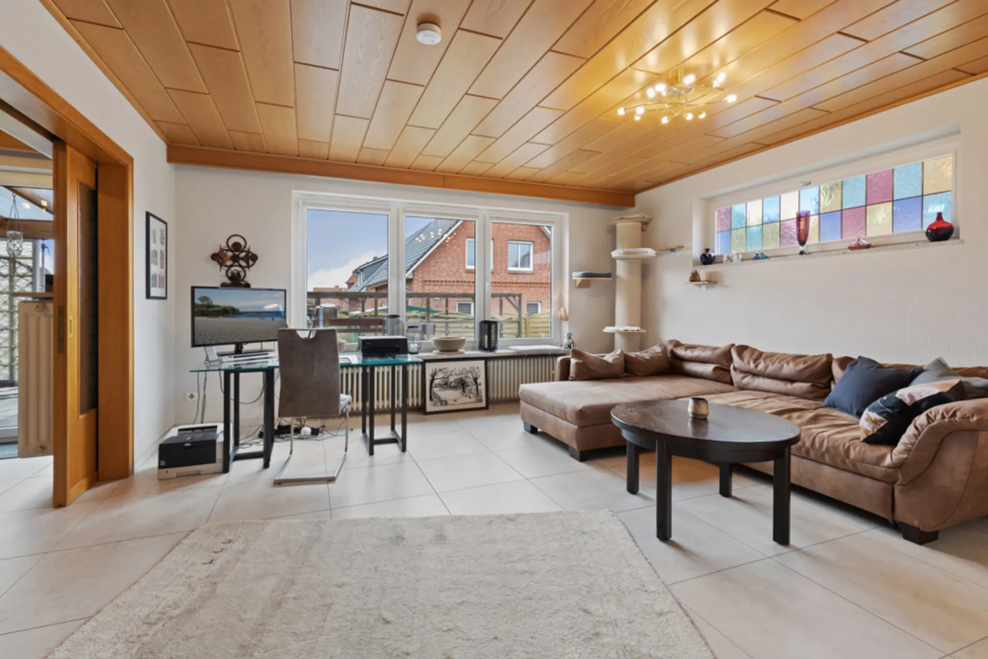 Helles Wohnzimmer mit Ecksofa, Couchtisch, Schreibtisch, Teppich und buntem Fenster.