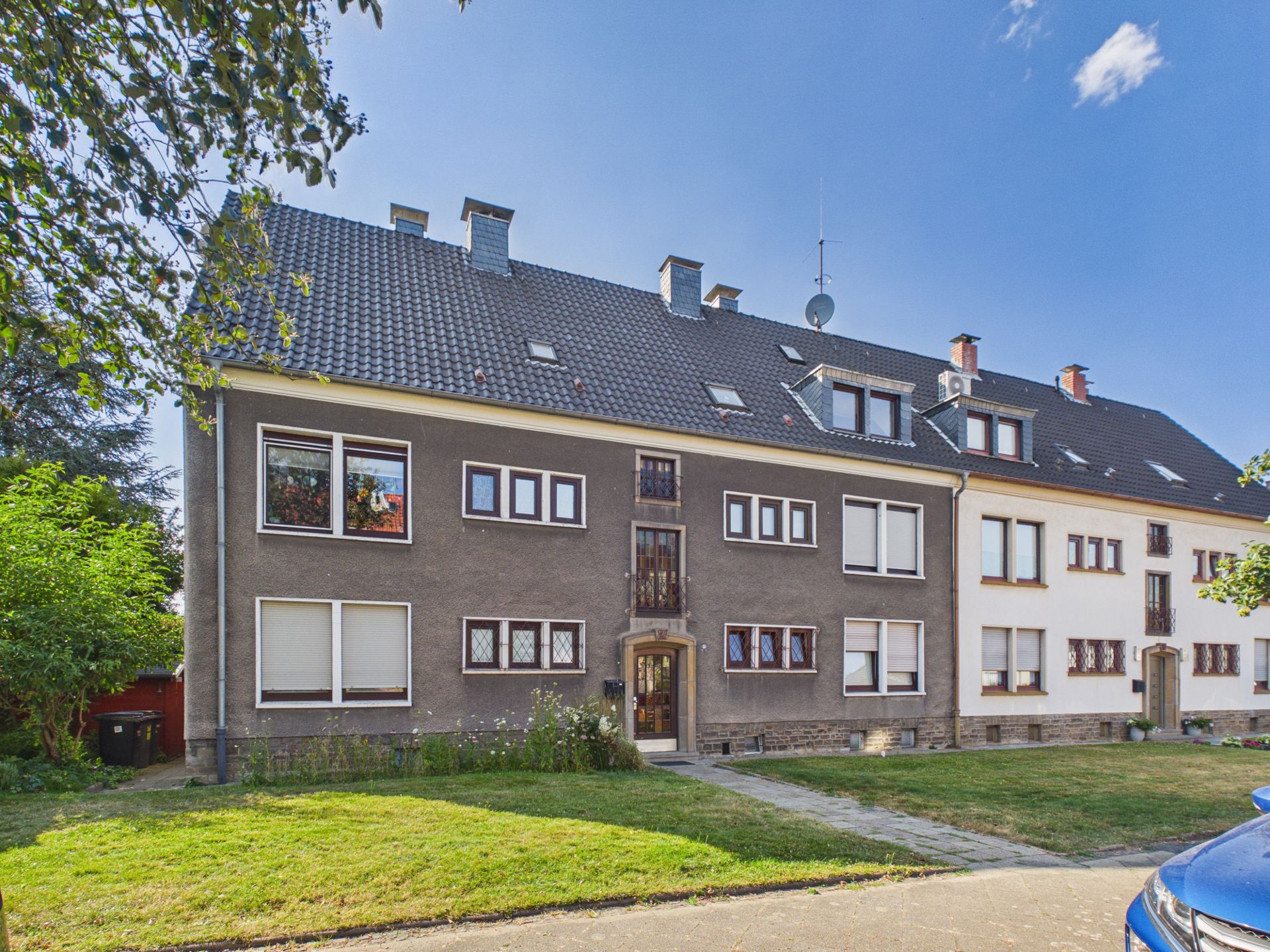 Mehrfamilienhaus mit dunkler Fassade, mehreren Fenstern und gepflegtem Vorgarten.