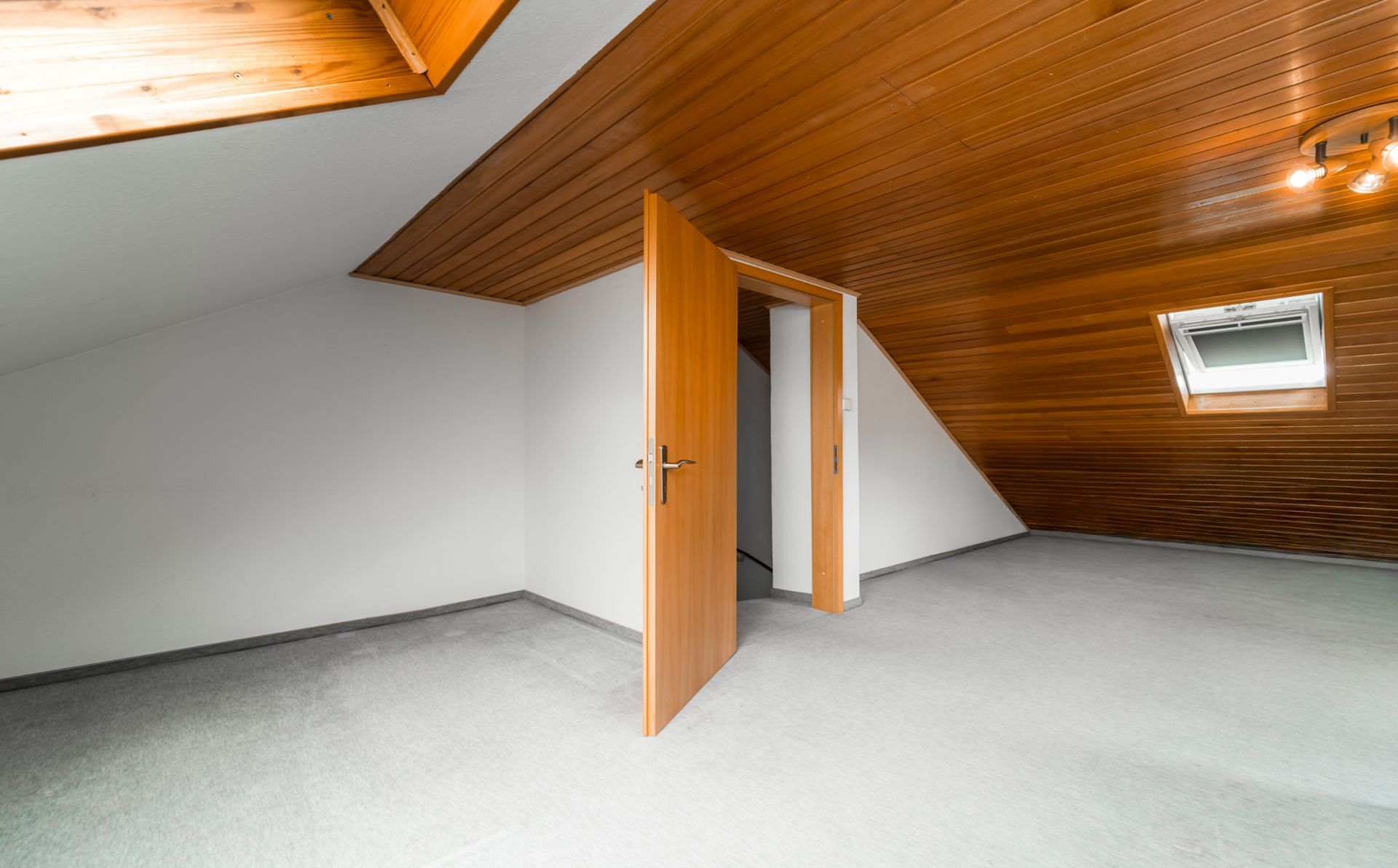Dachzimmer mit Holzdecke, Teppichboden, offenen Tür und Dachfenster.