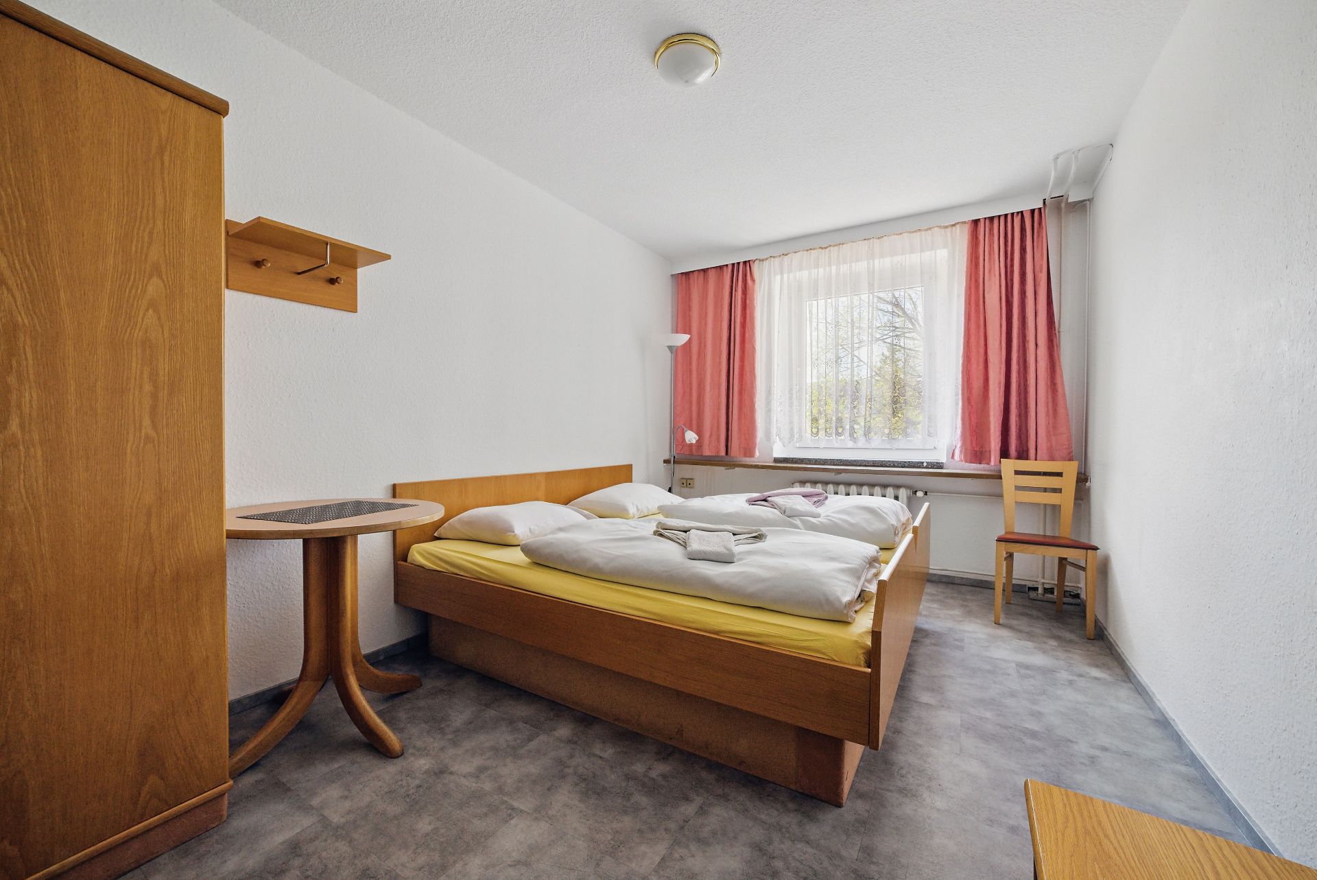 Ein einfach eingerichtetes Schlafzimmer mit Doppelbett, Holzmöbeln und Fenster mit roten Vorhängen.
