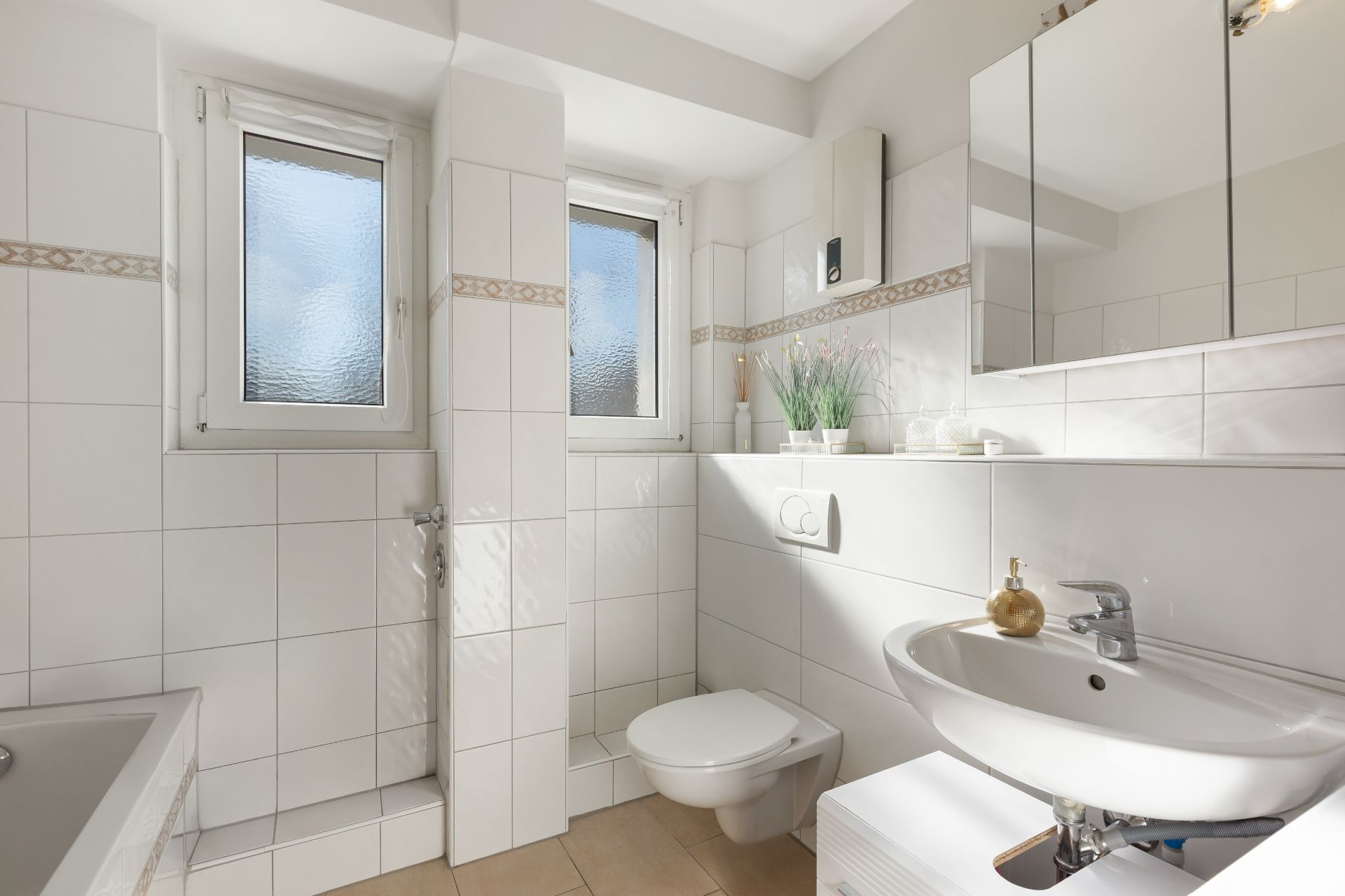 Helles Badezimmer mit weißen Fliesen, Toilette, Waschbecken, Spiegel und zwei Fenstern.