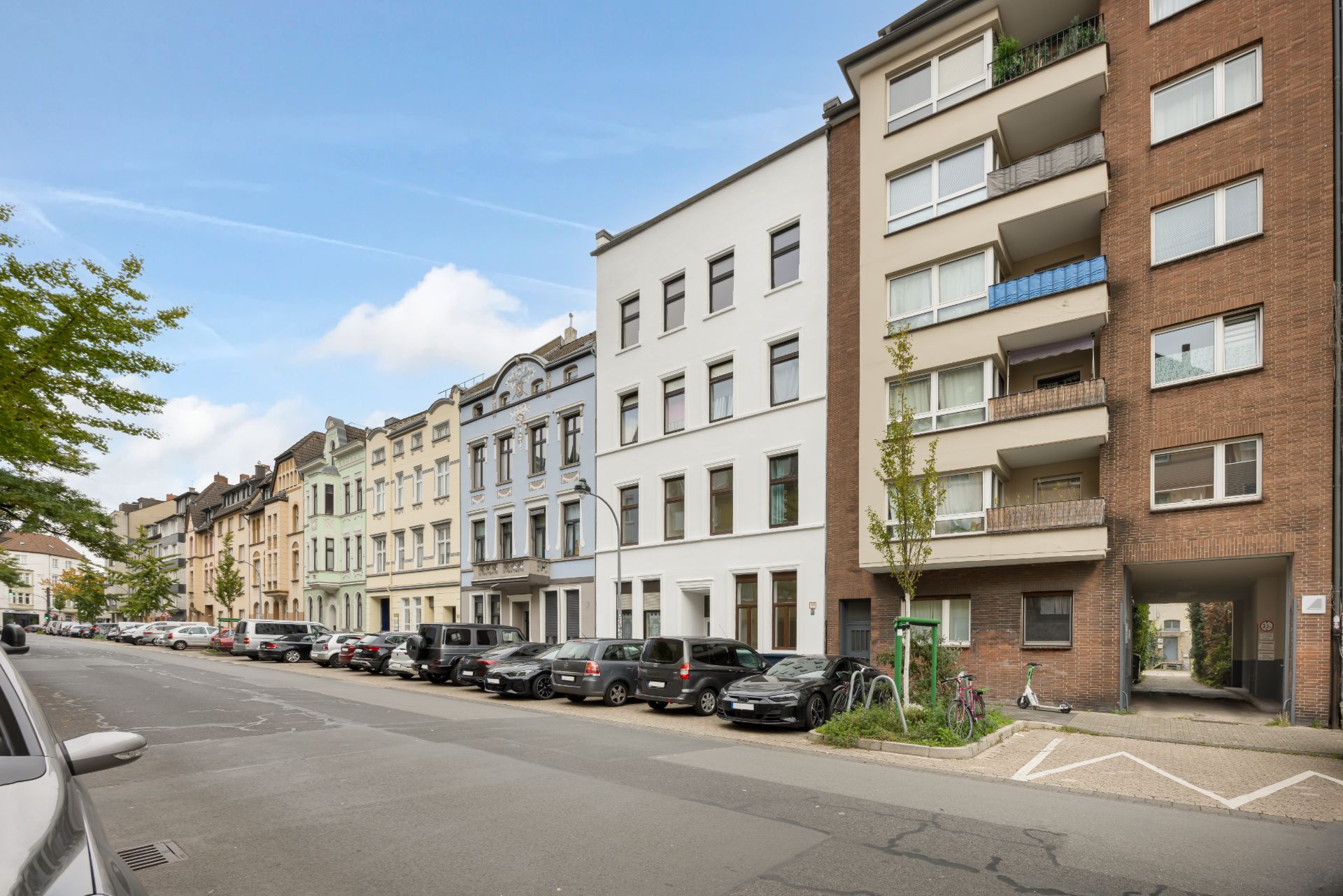 Straßenansicht mit mehreren Wohngebäuden und geparkten Autos am Straßenrand.