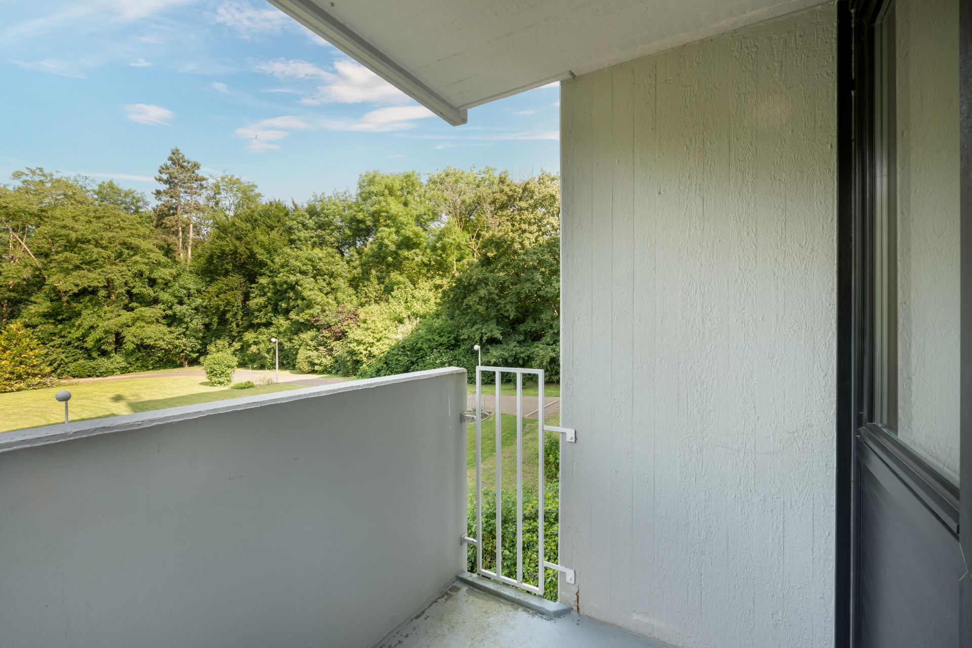 Kleiner Balkon mit weißem Geländer und Blick auf grüne Bäume und Rasenfläche.