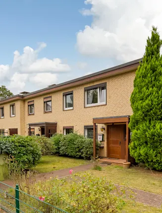 Reihenhaus mit gelber Fassade, Vorgarten, Hecken und großem Baum am Eingang.