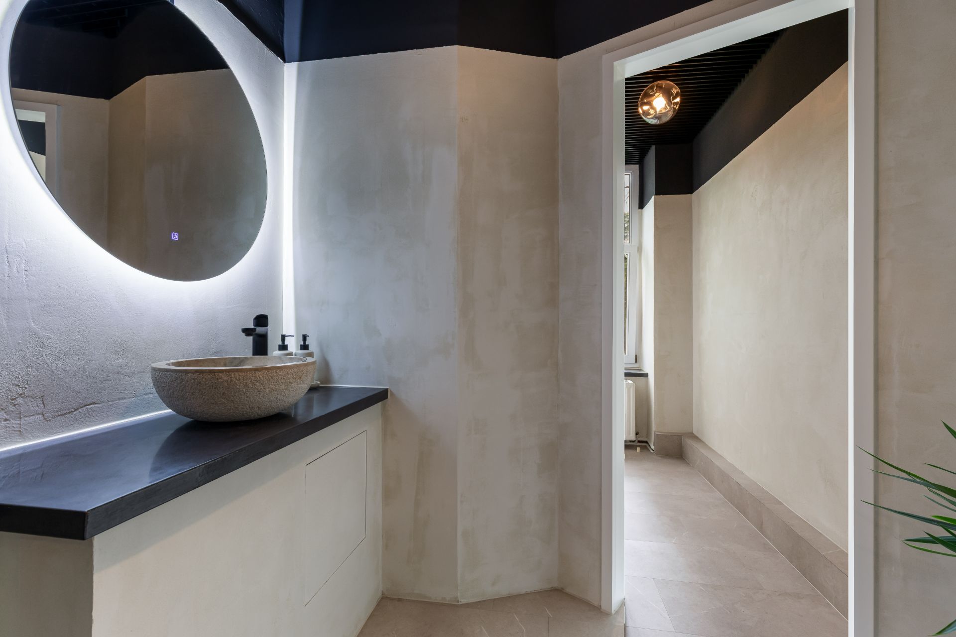 Modernes Badezimmer mit rundem Spiegel und Steinwaschbecken neben einer Tür zu einem Flur.