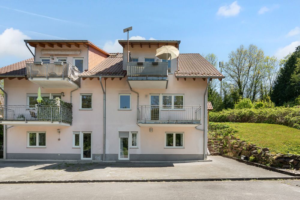Mehrfamilienhaus mit drei Balkonen und einem Garten im Hintergrund an einem sonnigen Tag.