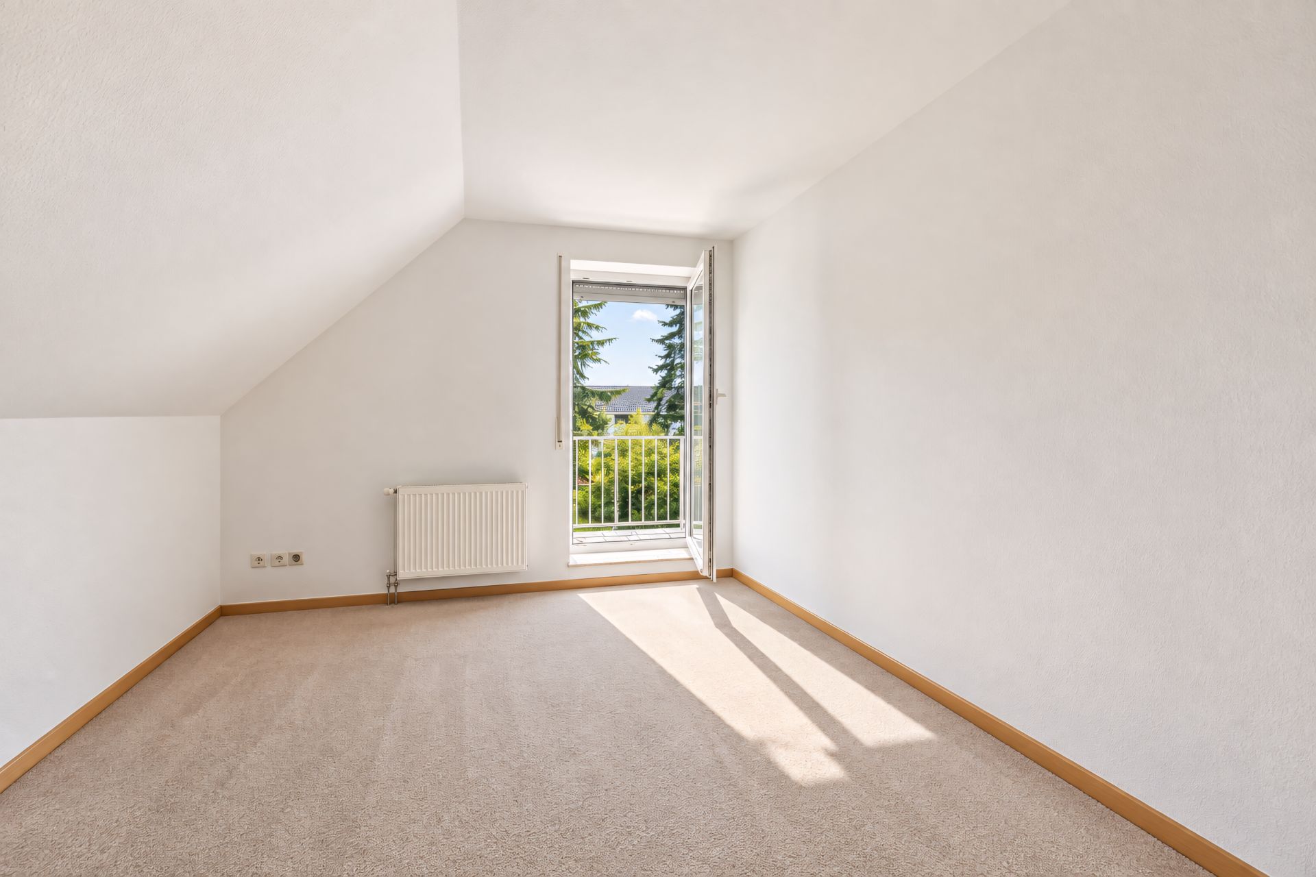 Helles, leeres Zimmer mit Teppichboden, schräger Decke und offenem Fenster mit Blick ins Grüne.