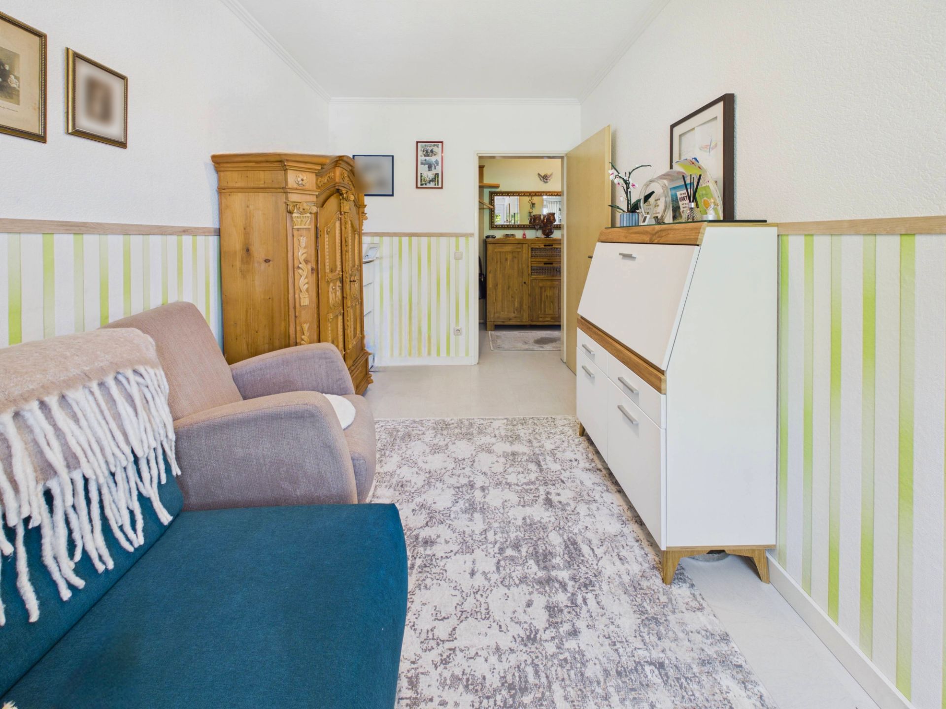 Helles Wohnzimmer mit Sofa, Sessel, Teppich, Schrank und Kommode, Wände mit grün-weißen Streifen.