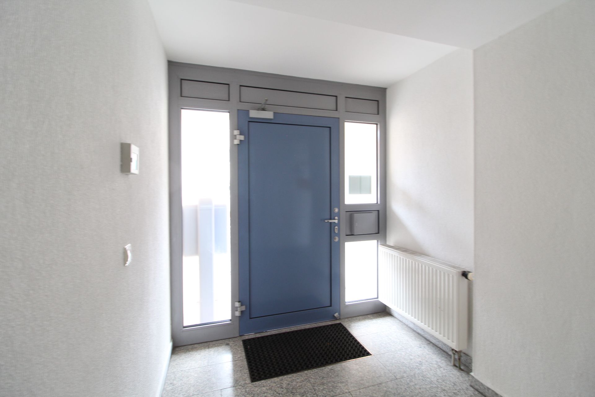 Heller Flur mit blauer Haustür, Fenstern, Heizkörper und Fußmatte.