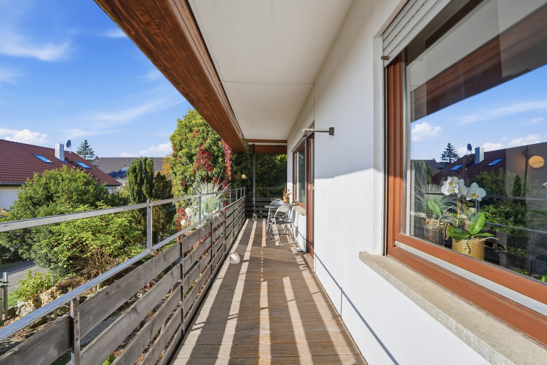 Langer Balkon mit Holzboden, Geländer und Blick auf Garten und Nachbarhäuser.