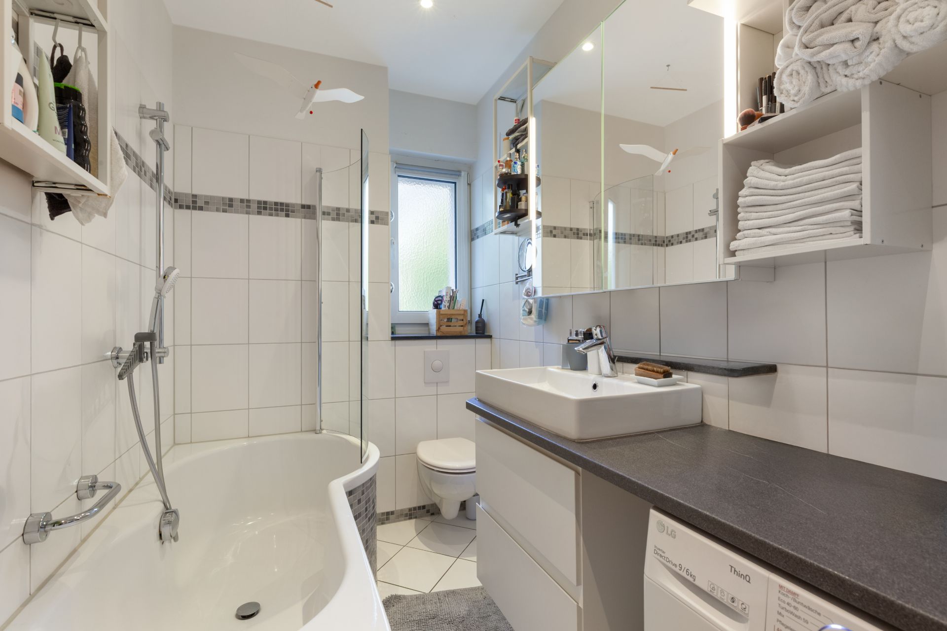 Modernes Badezimmer mit Badewanne, Waschbecken, WC, Spiegel und Regalen mit Handtüchern.