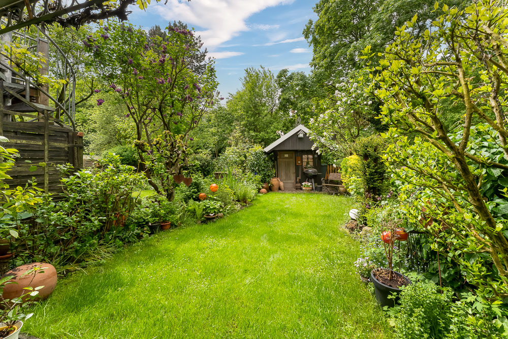 Grüner Garten mit Rasen, Blumen, Bäumen und einem kleinen Holzhaus im Hintergrund.