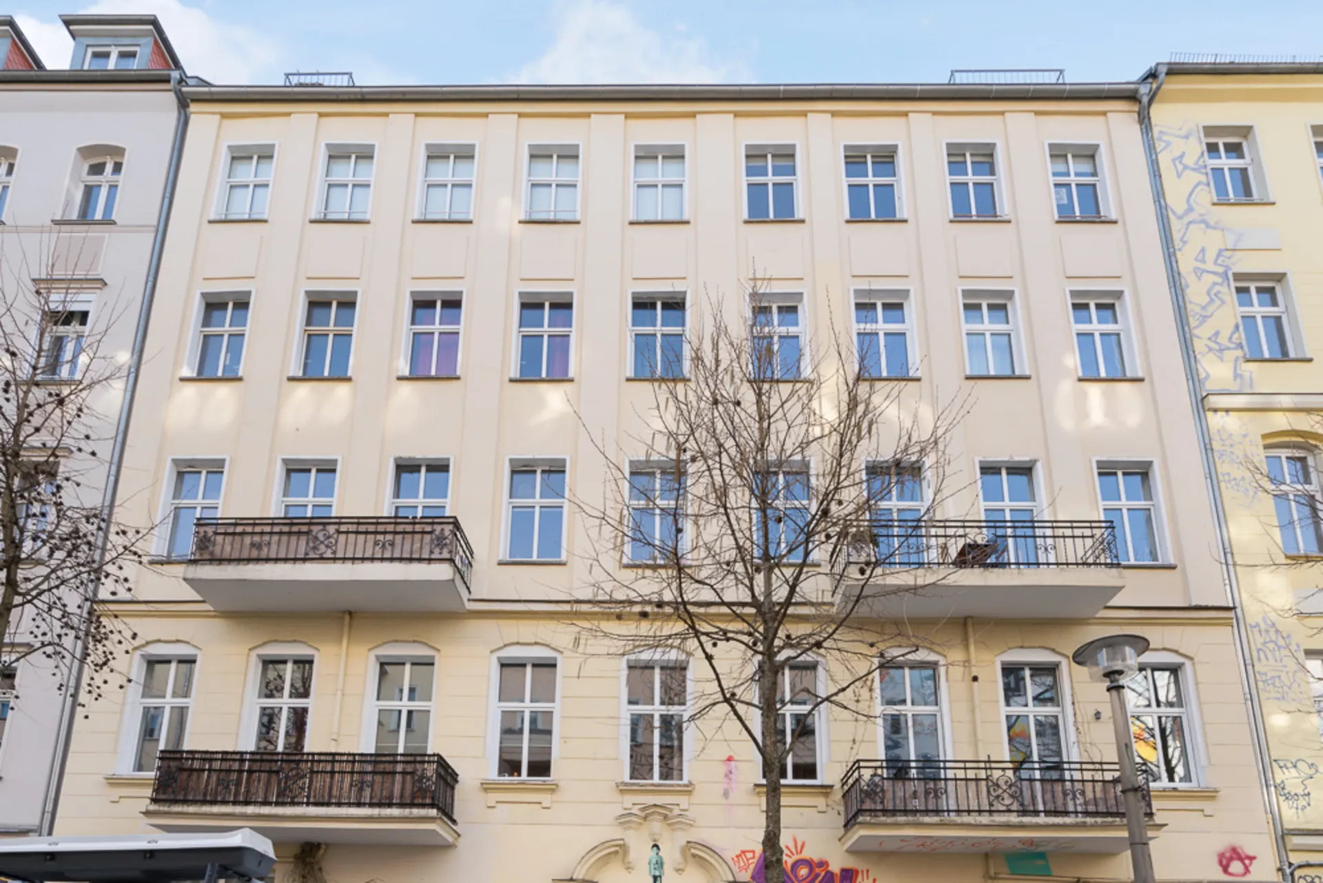 Vierstöckiges Wohngebäude mit mehreren Balkonen und Fenstern, Baum davor.