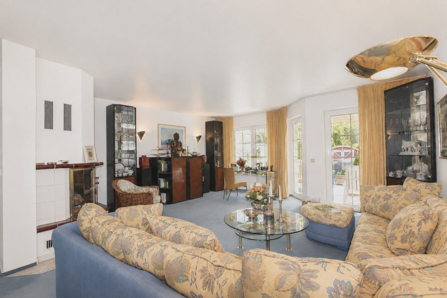Helles Wohnzimmer mit großer gemusterter Eckcouch, Glastisch und Fensterfront mit Blick nach draußen.