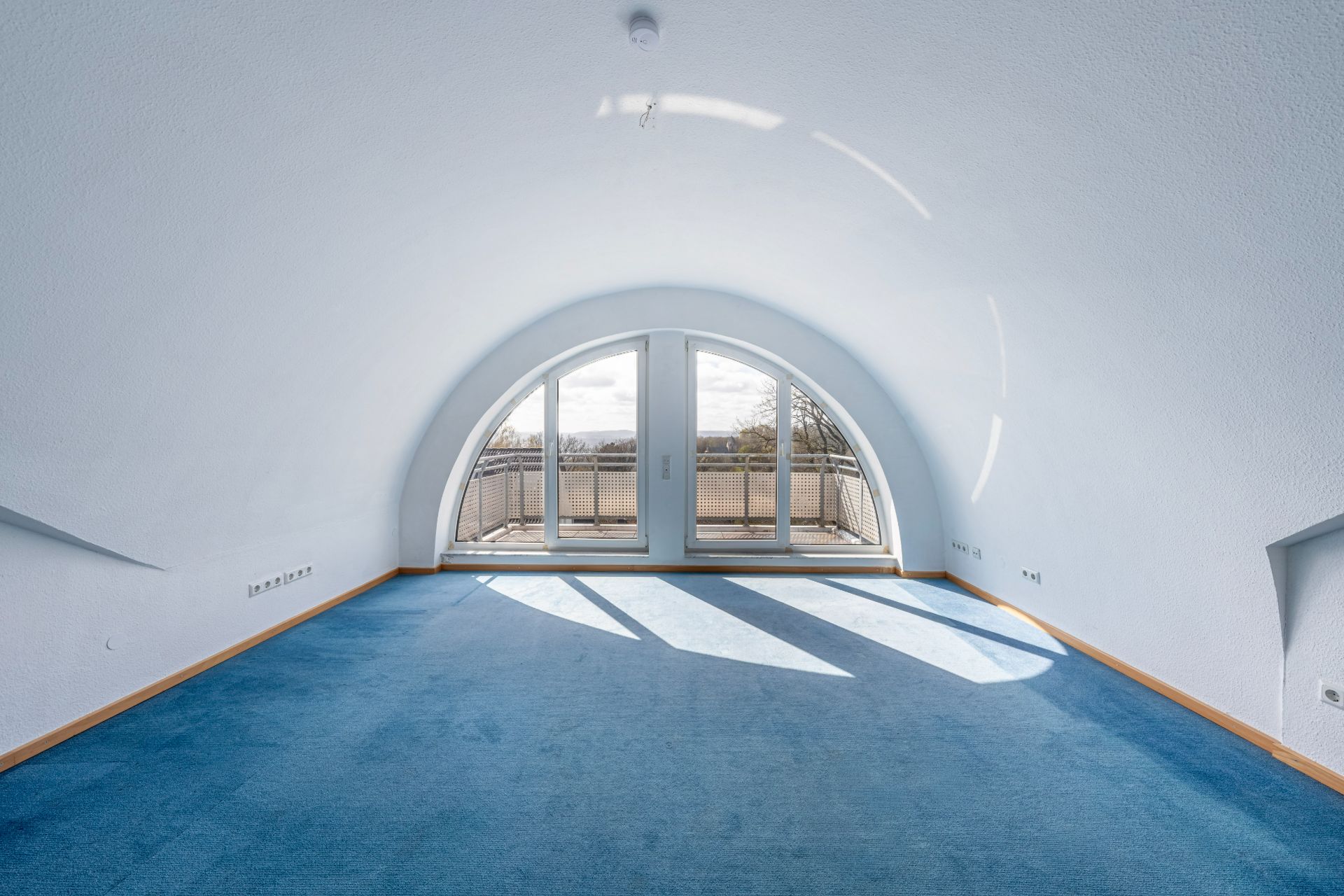 Helles Zimmer mit blauem Teppich, gewölbter Decke und großen Fenstern zu einem Balkon.