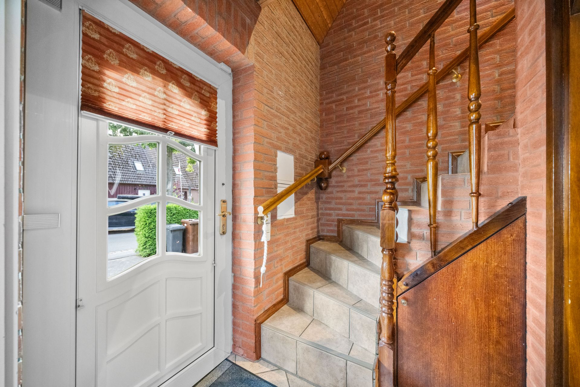 Hauseingang mit weißer Tür, Fenster und Treppe mit Holzgeländer, Wände aus rotem Backstein.