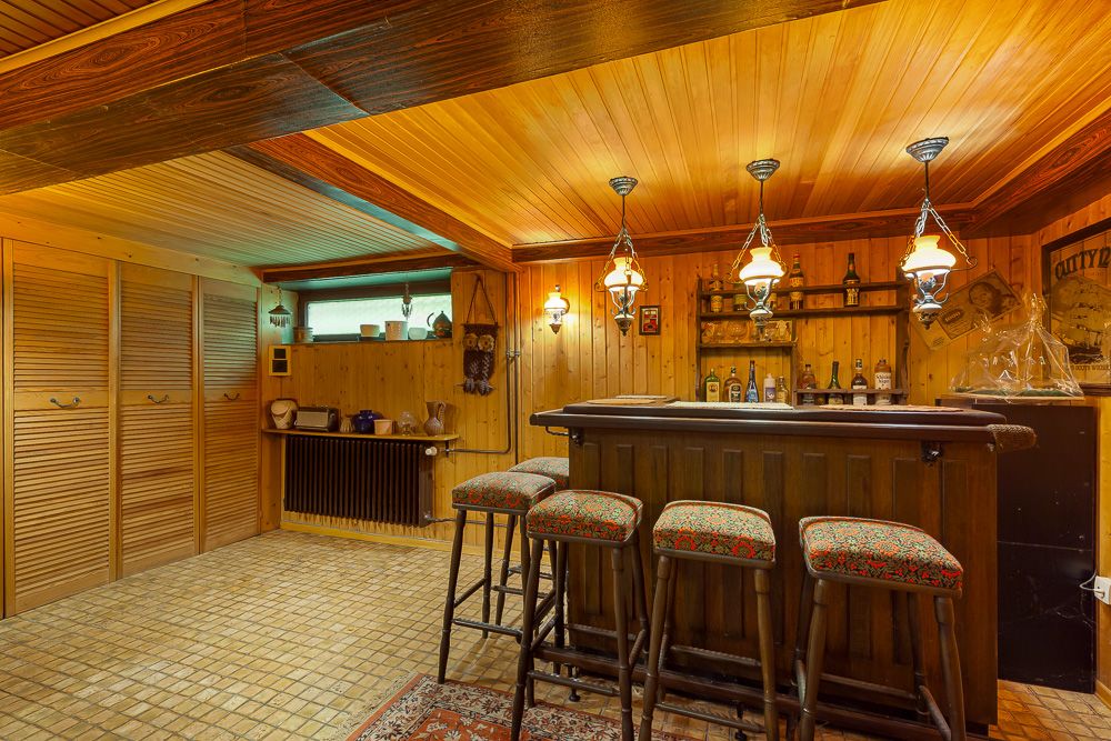 Gemütlicher Kellerraum mit Holzdecke, Hausbar, vier Barhockern und Regalen mit Flaschen.