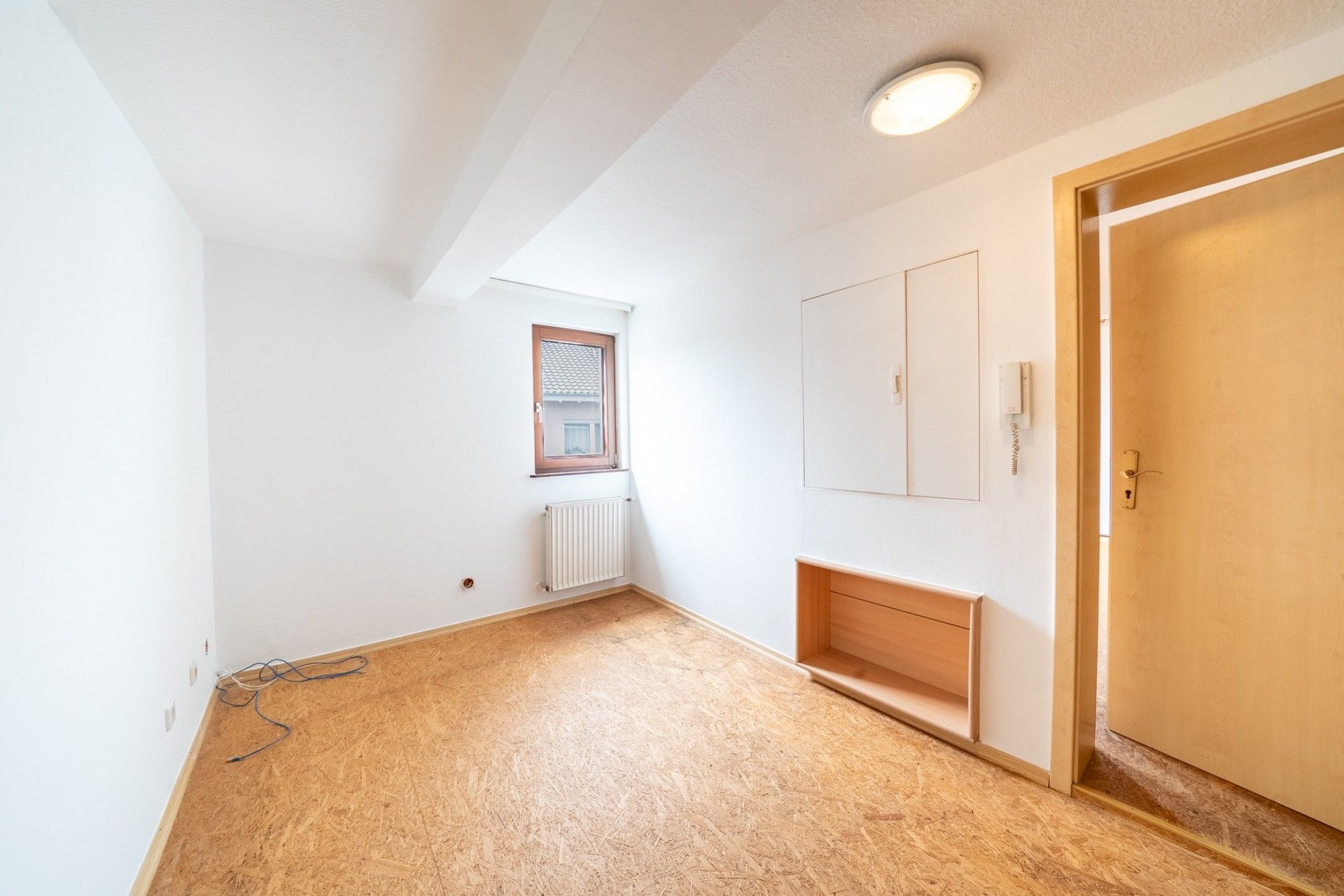 Leerer Raum mit hellem Boden, weißer Wand, kleinem Fenster und offener Tür.