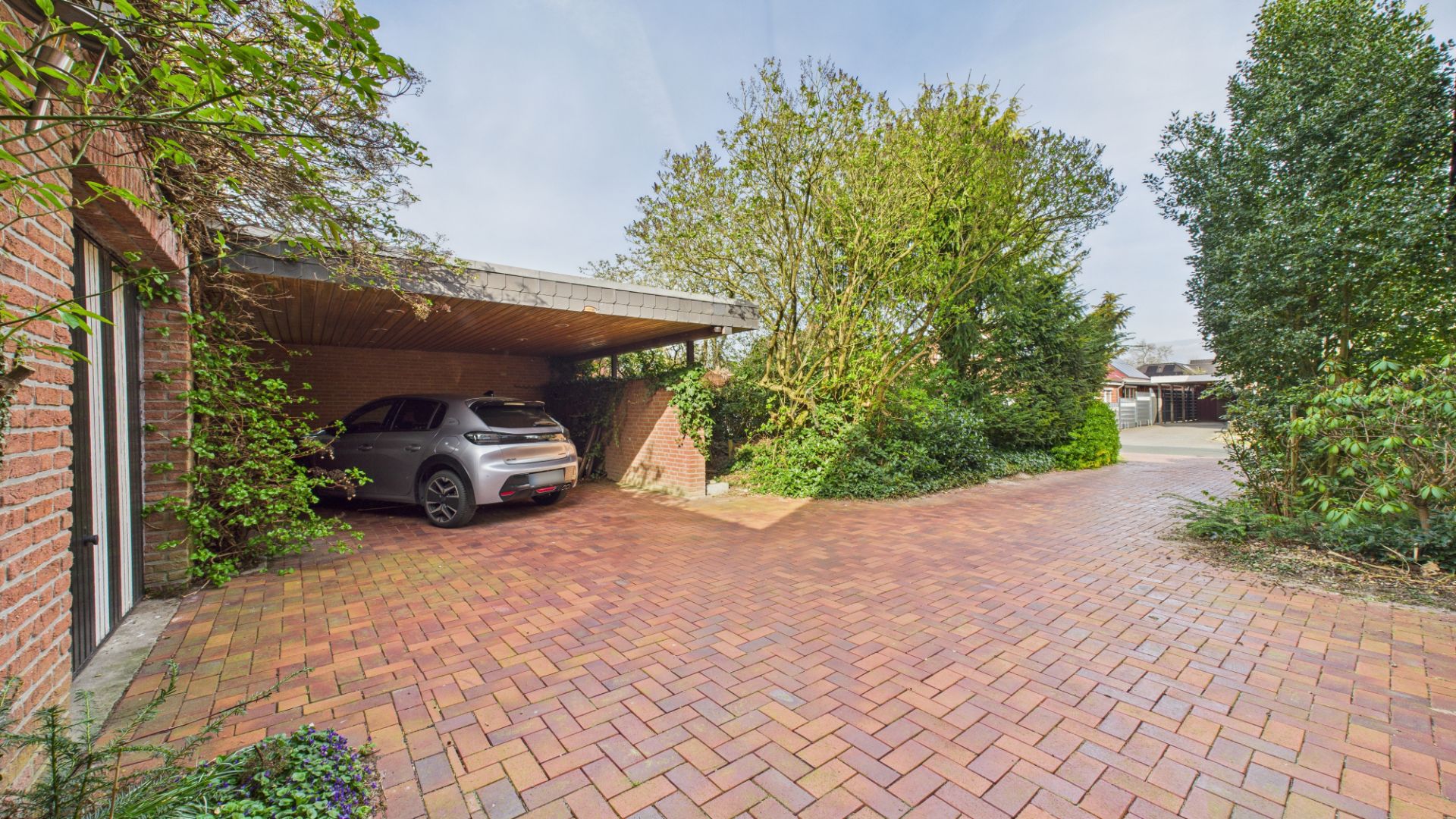 Pflasterstein-Auffahrt mit Carport und geparktem Auto, umgeben von Bäumen und Sträuchern.