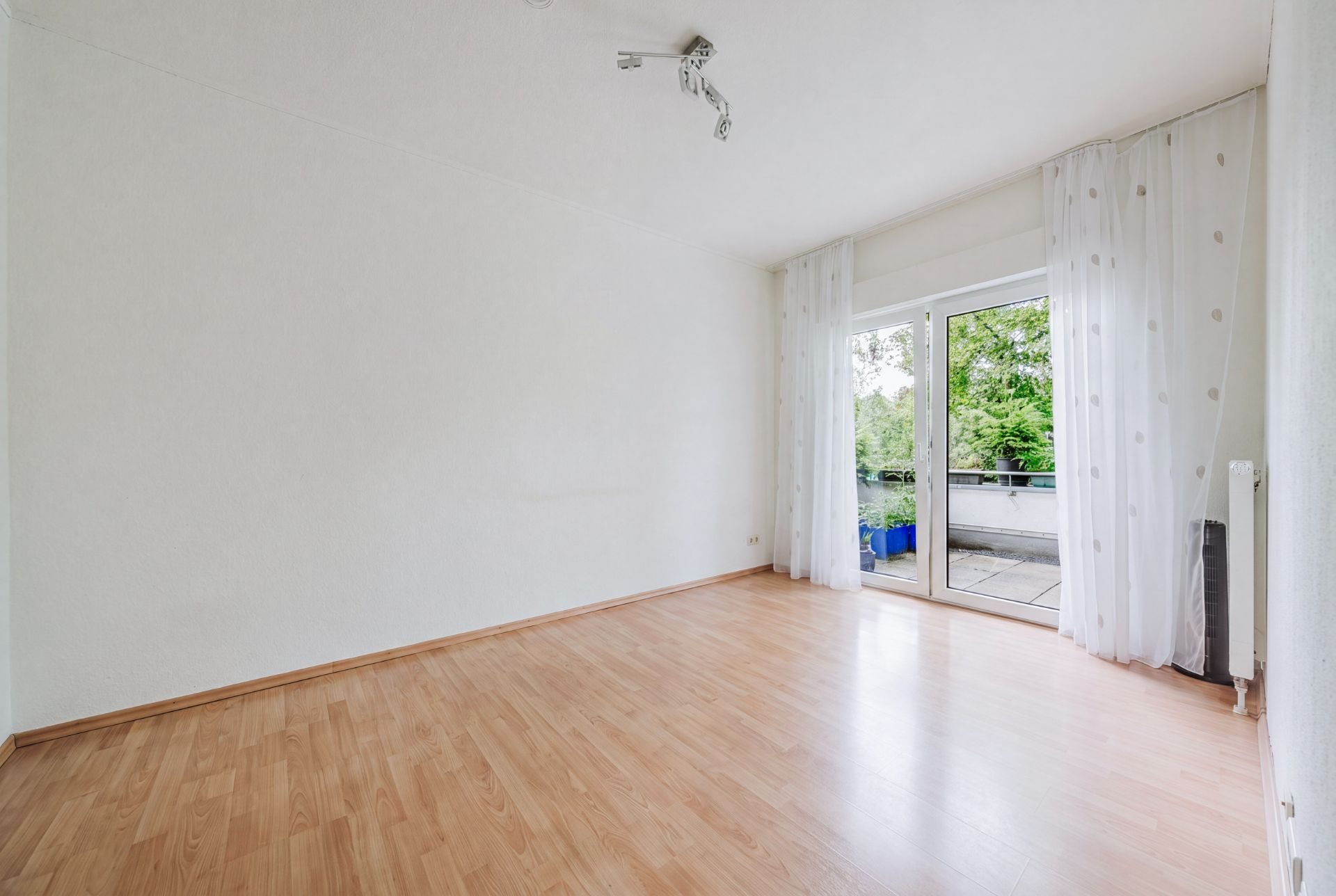 Helles, leeres Zimmer mit Holzfußboden und großen Fenstertüren zum Balkon.