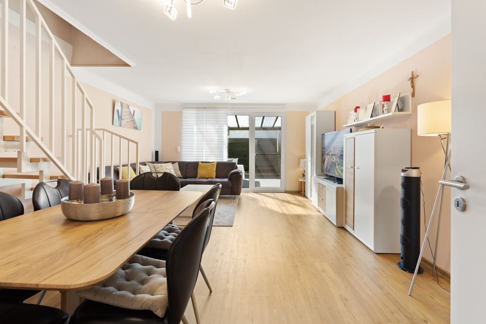 Helles Wohnzimmer mit Esstisch, Sofa, Fernseher und Treppe, Holzboden und großen Fenstern.