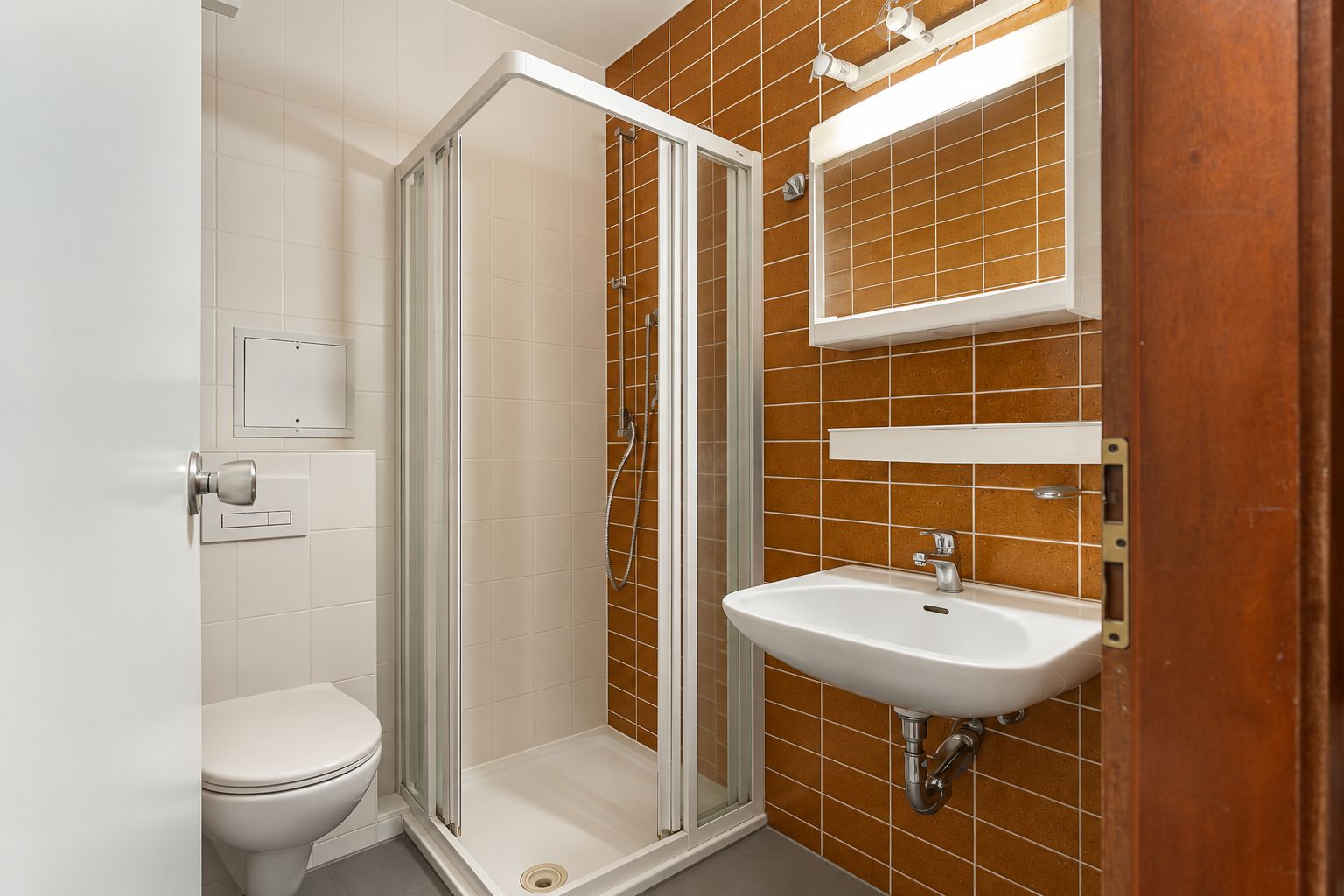 Kleines Badezimmer mit Dusche, WC, Waschbecken und braunen Fliesen an einer Wand.