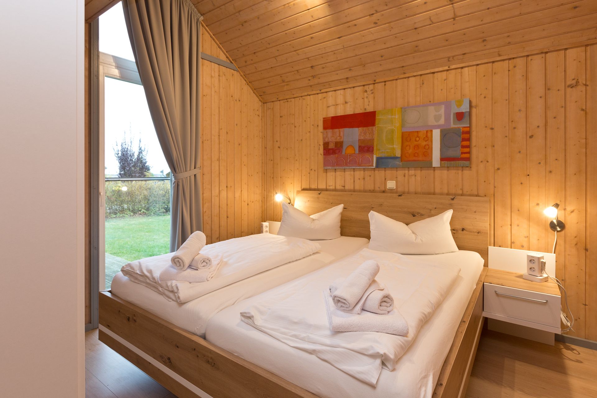 Schlafzimmer mit Doppelbett, Holzverkleidung, Kunst an der Wand und großem Fenster mit Blick ins Grüne.