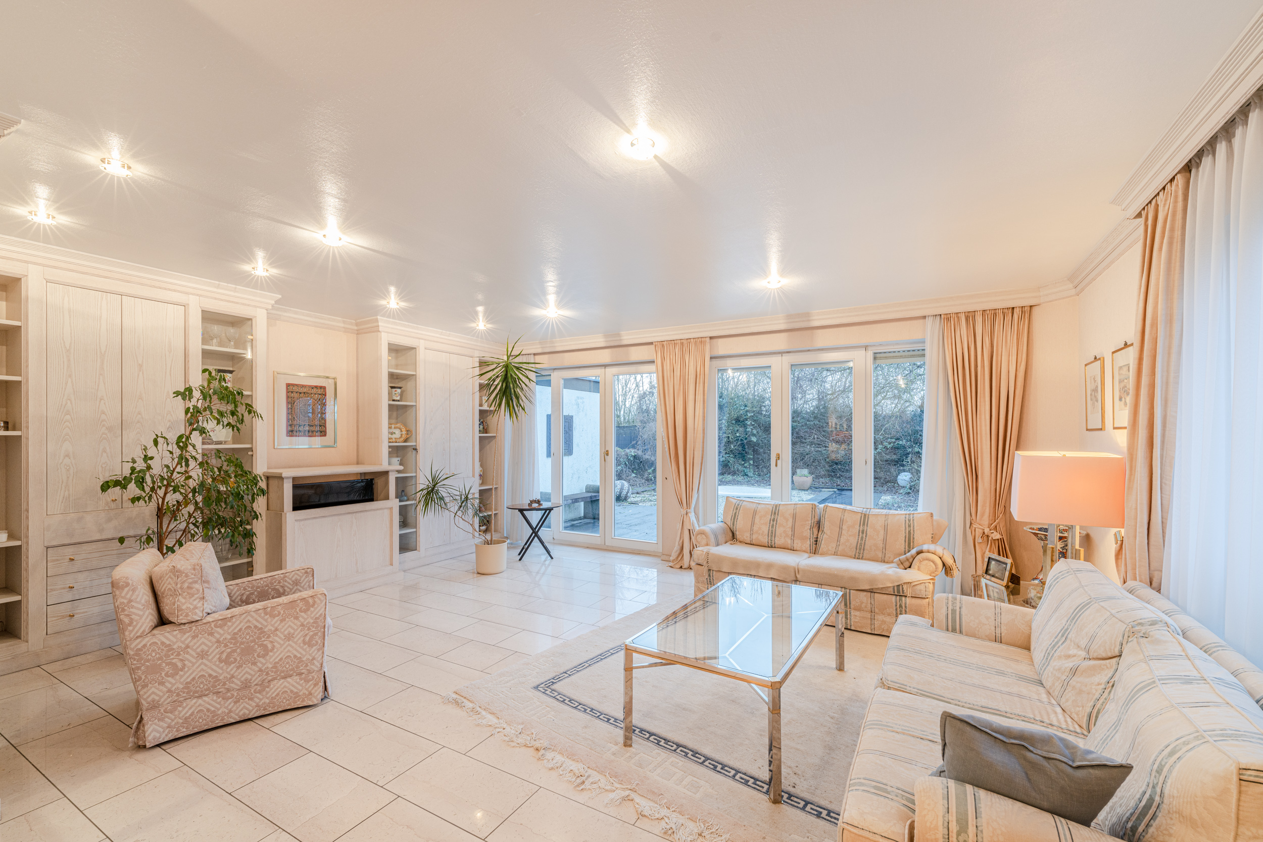 Helles Wohnzimmer mit großen Fenstern, zwei Sofas, Sessel, Glastisch und Pflanzen.