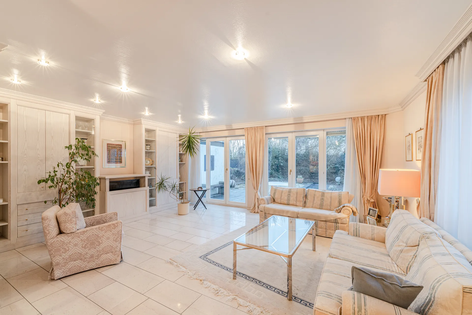 Helles Wohnzimmer mit großen Fenstern, zwei Sofas, Sessel, Glastisch und Pflanzen.