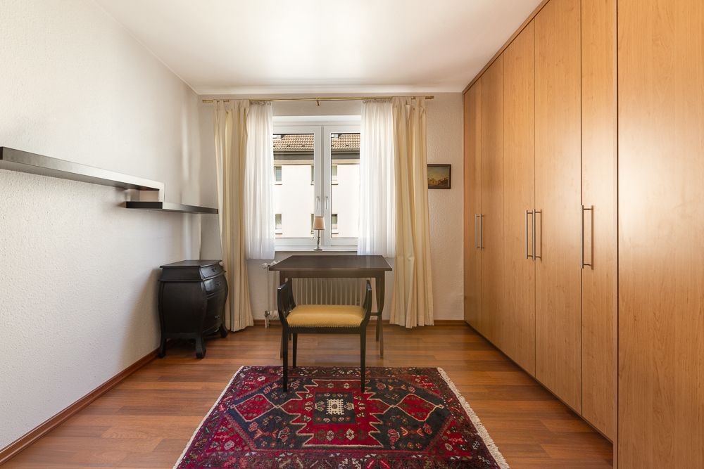 Helles Zimmer mit großem Schrank, Schreibtisch, Stuhl, Teppich und Wandregal.