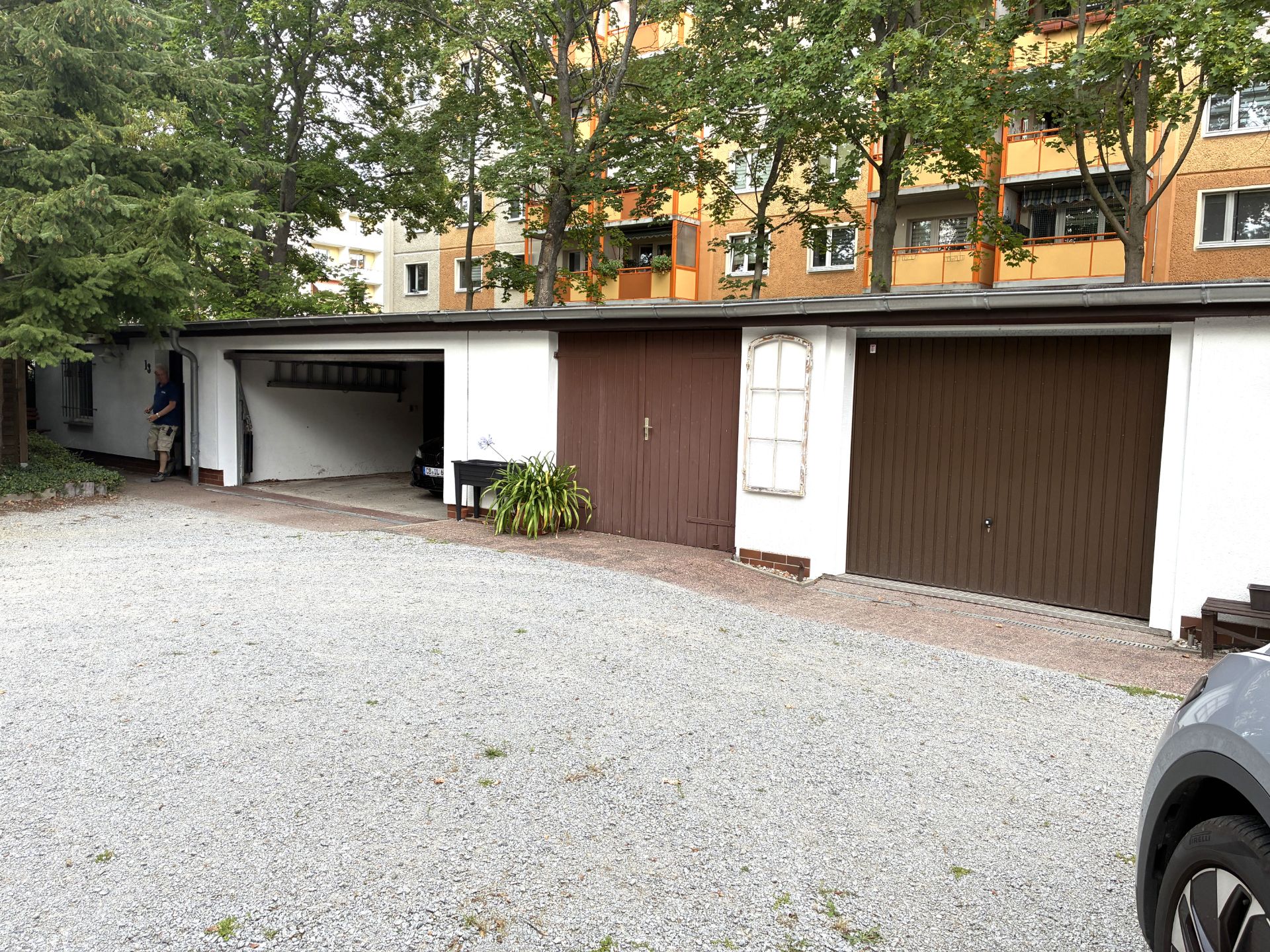 Drei Garagen mit braunen Türen und ein offenes Carport auf einem Hof, Bäume und Wohnhaus im Hintergrund.