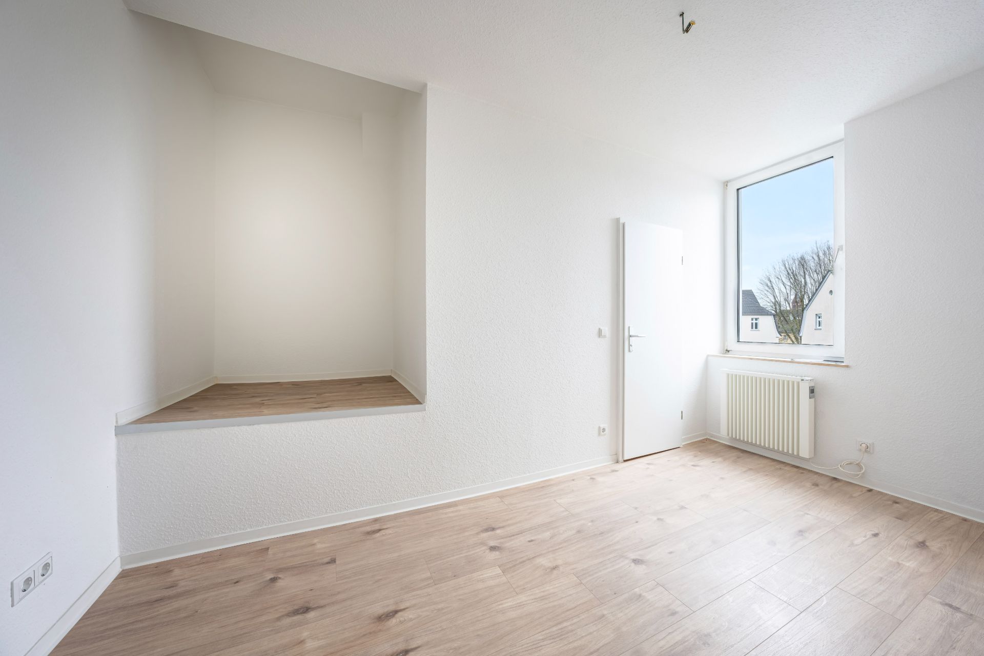 Helles leeres Zimmer mit Holzboden, Fenster, Tür und erhöhtem Podestbereich.