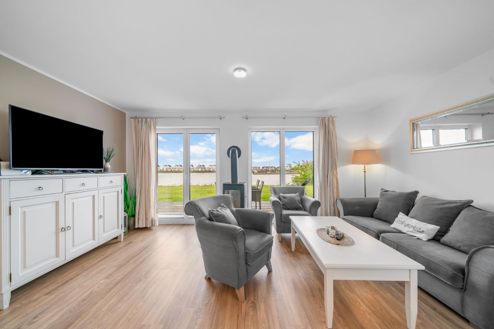 Helles Wohnzimmer mit Sofa, zwei Sesseln, weißem Tisch und großem Fenster zum Garten.